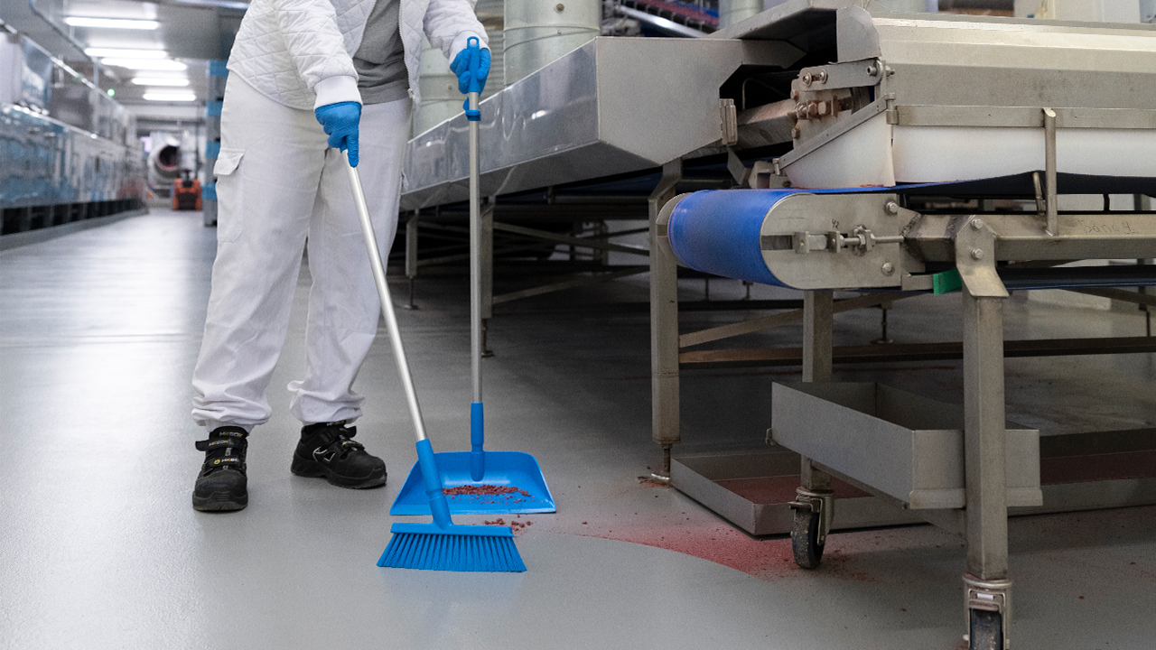 Industrial setting with a worker using a blue Vikan dustpan and broom for cleaning food processing residue.