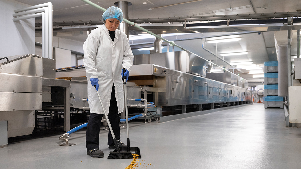 Worker in food production using a black Vikan dustpan and broom set to maintain cleanliness.
